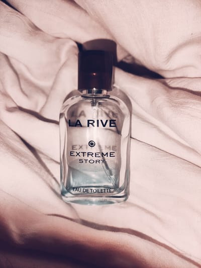 The image shows a clear glass bottle of "LA RIVE EXTREME STORY EAU DE TOILETTE" perfume. The bottle has a dark brown cap and is positioned on a wrinkled, light pink fabric, possibly a blanket or sheet. The lighting is soft, casting subtle shadows. The background is blurred, with the focus on the perfume bottle. There is no indication of the city of Murska Sobota, Slovenia, in the image.