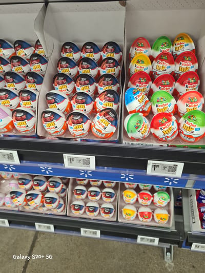 The image depicts a display of Kinder Joy chocolates in a retail store, likely a Walmart based on the blue shelves and pricing tags. The chocolates are arranged in rows within cardboard dividers. There are two distinct types of Kinder Joy shown: one featuring characters from "Stranger Things" and another with "Super Mario" characters. The "Stranger Things" themed chocolates have red, white, and blue wrappers with "Funko" and "Stranger Things" branding. The "Super Mario" chocolates have various colored wrappers, including red, green, blue, and yellow, with "Super Mario" branding. Some of the "Super Mario" chocolates are also visible on a lower shelf. Pricing tags for the Kinder Joy Variety are visible in the foreground, indicating a price of $1.97. The background shows more shelves and products, suggesting a well-stocked retail environment. The overall scene is brightly lit and appears to be a product display within a store. The timestamp "Galaxy S20+ 5G" is visible in the bottom left corner of the image. This scene is likely located in Woodburn, Oregon, USA.