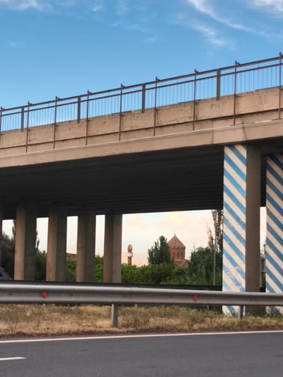 The image shows an overpass in Artashat, Armenia. The overpass is made of concrete and has a metal railing on top. The pillars supporting the overpass are decorated with blue and white diagonal stripes. In the background, a red-brick church with a conical roof can be seen amongst trees. The sky is blue with a few white clouds. In the foreground, there is a road with a metal guardrail. The overall scene suggests a rural or suburban area.