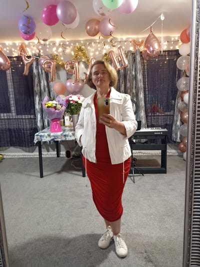 A woman is standing in front of a mirror in a room decorated for a celebration. She is wearing a red ruched dress and a white jacket, along with white sneakers. She is holding a gold-colored smartphone in her right hand, taking a selfie. The background shows a table with a bouquet of flowers, balloons, and string lights, indicating a festive occasion. A keyboard is visible in the reflection of the mirror. The room has carpeted floors and patterned curtains. The city of Annagry, Ireland is mentioned, but there is no specific visual information in the image to confirm this location.