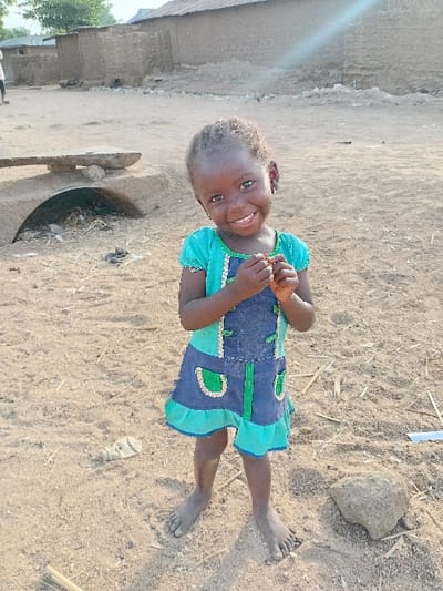 An image taken in Nata-Ala, Nigeria, depicts a young Black child, approximately 2-4 years old, standing barefoot and centered in a dry, sandy outdoor environment. The child wears a short-sleeved dress with a teal/light blue top and a dark blue bottom half featuring green fabric accents and light trim. The child is smiling broadly at the camera, with hands clasped together near the chin. The ground beneath the child is light brown and dusty, with scattered debris including small sticks, dry plant matter, and a crumpled white plastic bottle to the left. A small gray rock is positioned to the child's right. In the mid-ground to the left, a low, arched mud or concrete structure is visible, topped with a wooden plank and containing debris within its opening. The background features several simple, single-story buildings constructed of mud or adobe, with rough textures and indistinct roofing, indicative of a rural or semi-urban settlement. Bright sunlight is present, causing a lens flare in the upper right portion of the frame.