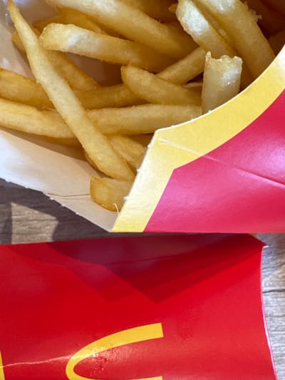 The image depicts a close-up of McDonald's french fries in a partially opened red and yellow paper container, resting on a light-colored, textured surface, possibly a wooden table. Another closed McDonald's french fry container is positioned to the right of the open one. The location in Lipomo, Italy is not explicitly stated or visually discernible from the image itself.