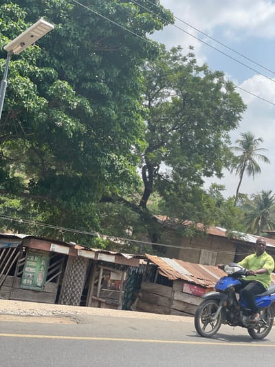 The image depicts a roadside scene in Ozuri, Nigeria, under daylight conditions. In the foreground, a paved asphalt road with a solid yellow center line runs horizontally. An adult male, with dark skin and a receding hairline, operates a blue and dark-colored motorcycle from right to left across the frame. He wears a light green short-sleeved polo shirt, dark trousers, and sandals. His hands are positioned on the handlebars.
Behind the road, a row of approximately four single-story commercial structures or stalls are visible. These structures feature weathered wooden walls and rusted corrugated metal roofs of varying heights and angles. One stall on the far left displays a poster with the word "Honourable" and an image resembling a beer bottle. Another stall shows patterned fabric covering part of its opening, while a third reveals shelves with items, possibly beverages.
Above and behind these structures, a dense canopy of various trees dominates the midground and background. A large tree with broad, dark green leaves occupies the left side of the frame, extending upwards. Further right and behind, other trees with lighter green foliage are present, along with at least two slender palm trees visible on the far right. A modern, rectangular solar-powered streetlight on a pole extends upwards on the far left. Utility lines are visible horizontally across the upper portion of the frame. The sky above is partially cloudy.