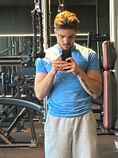 An image captured in a gymnasium in Jestetten, Germany, depicts a male subject taking a mirror selfie. The subject, a Caucasian male with light brown hair styled upwards and a short beard, stands facing forward, looking down at a dark-colored smartphone held in both hands. He is wearing a light blue short-sleeved t-shirt and light gray sweatpants. Script tattooing is visible on his inner left forearm. The background, visible as a reflection in the mirror, shows various gym equipment, including a cable machine with a rope attachment, weight stacks, and upholstered pads on other machines. Overhead spotlights are also reflected, illuminating the scene.