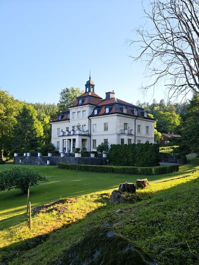 A stately, pale yellow mansion with a red tile roof sits nestled amidst lush green trees and manicured lawns. The mansion features a central tower with a dark dome, multiple windows, and balconies. White columns support a porch on the front facade. A stone retaining wall with decorative white railings lines the elevated front yard, accessed by stone steps.
To the right of the mansion, a smaller, red-colored building with a red roof is partially visible, suggesting a secondary structure on the property. The surrounding landscape is verdant, with a gently sloping lawn bathed in the warm glow of what appears to be late afternoon or early morning sunlight, casting long shadows across the grass. Tall, mature trees and dense foliage surround the buildings, creating a sense of seclusion and natural beauty. Bare branches of a large tree are silhouetted against a clear, bright blue sky in the upper right portion of the image.
In the foreground, several large rocks and tree stumps are scattered on the grassy hillside. A wooden fence post stands in the middle ground. The scene conveys a sense of tranquility and peacefulness, characteristic of a rural estate in a temperate climate. There is no visible text in the image. The location context provided is Skriketorp, Sweden.
