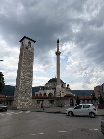 The image depicts the Gazi Ali Pasha Mosque and the adjacent Clock Tower in Pljevlja, Montenegro. The sky is overcast with gray clouds. A tall, stone clock tower with a dark roof stands to the left of the frame, displaying a round clock face. To the right of the tower, the mosque features a dome and a slender minaret. A white car is parked on the right side of the street, and a black car is driving on the left. A man wearing a red shirt and blue pants walks across a crosswalk in the middle of the street. Buildings, trees, and a hillside with trees are visible in the background. The street in the foreground is paved with asphalt and has white lane markings.