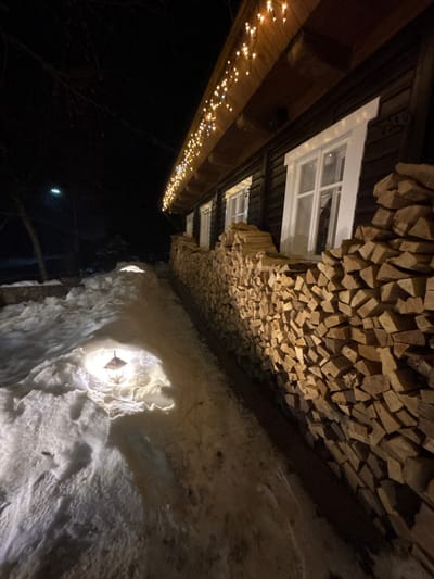 The image captures an outdoor night scene featuring a building, a wall of stacked firewood, and an illuminated snow or ice wall. The perspective is tilted, making the building appear to be on the left side of the frame, running vertically.

The structure on the left appears to be a building, likely made of dark wood planks or logs. Several white-framed windows are visible on its facade. Below a prominent wooden eave or beam that runs along the building, numerous small, warm-toned string lights are strung, casting a festive glow downwards into the dark, unseen area below. These lights appear to be clustered or star-shaped.

Adjacent to the building's facade, towards the right, is a substantial wall constructed from neatly stacked pieces of light-brown, irregularly cut firewood. The stacking creates a dense, textured surface for this wall, which appears to rise alongside the building.

Further to the right, and occupying a significant portion of the lower and middle-right of the frame, is a highly textured wall composed of what appears to be snow or ice. This surface is uneven and rugged, exhibiting shades of bright white and light brown. Several bright light sources are embedded within or positioned very close to this snow/ice wall, illuminating its contours dramatically. One prominent light fixture, possibly a metal lantern or sconce, is clearly visible within a carved-out section of this wall, casting a strong, warm white light that highlights the crystalline and rough textures of the snow/ice and creates pronounced shadows. The overall effect on this section is grotto-like or cave-like.

The background to the far left is dark, indicating nighttime, with faint suggestions of tree branches or distant objects. A very dim, small light source can also be discerned in the dark expanse. The overall lighting suggests a cold weather setting, possibly winter, with a festive and somewhat magical ambiance created by the contrasting warm and cool lights. No text is visible in the image.