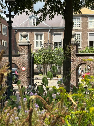 The image captures a view through a lush garden towards a historic building and a gateway in Roermond, Netherlands. In the foreground, out-of-focus wild grasses and flowering plants with purple and yellow blooms create a natural screen. Beyond this, a tall, dark lamppost stands to the left, partially obscuring the view.

A brick gatehouse with an arched opening is central to the mid-ground. A circular red and white sign is attached to the brick pillar on the left side of the gatehouse. Through the archway, a dark-colored car is visible. The gateway itself features ornate wrought-iron fencing.

Behind the gatehouse, a stately brick building with multiple windows and grey awnings is visible. The building has a pitched roof with dormer windows. The architectural style suggests historical significance, typical of older European cities like Roermond.

In the background, through another arched opening in the brickwork on the right, two people are partially visible. One, wearing a dark shirt, is pushing a red cart. Another person with grey hair and wearing a striped shirt is seated.

The overall scene depicts a serene urban environment, where nature and historical architecture coexist. The image appears to be taken on a sunny day, with dappled light filtering through the leaves of a tree in the upper right portion of the frame.