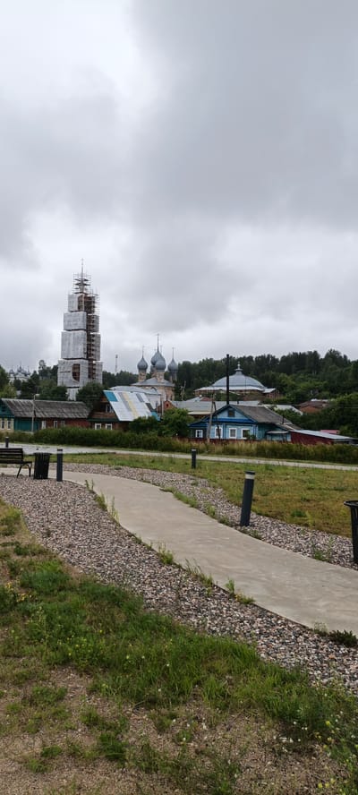 In Yuryevets, Russia, under an overcast sky, a paved pathway meanders through a landscape of mixed gravel and grass. To the left, a dark park bench and a cylindrical trash receptacle sit near the path's beginning. In the mid-ground, a row of village houses, constructed from wood and featuring colorful tin roofs, stretches across the scene. Behind the houses, a complex of church buildings with distinctive blue onion domes stands, and further to the left, a tall, bell tower is partially encased in scaffolding and white sheeting, suggesting ongoing construction or renovation. A dense line of green trees forms a backdrop to the settlement, with a gentle hill visible beyond. The overall impression is one of a rural village with historical architecture, actively undergoing preservation.