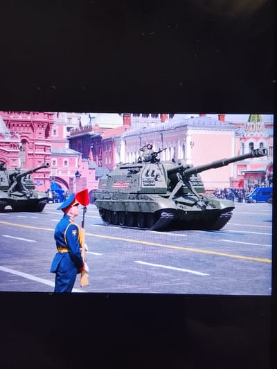 In Safonovo, Russia, a military parade is underway. A soldier in a blue uniform with gold trim and a red hat stands at attention holding a rifle with a red flag attached to it. Behind the soldier, a large, green self-propelled howitzer is driving down the street. The howitzer is a 2S19 Msta-S. In the background, there are traditional Russian buildings with red brick facades and ornate architecture. Another military vehicle, a tank with a red "Z" symbol on its side, is also visible in the parade. A blue van is parked on the side of the road, and a crowd of spectators can be seen in the distance. The sky is clear and the sun is shining, casting shadows on the street.