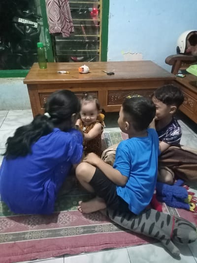 Four children are gathered on a patterned rug indoors, likely in a living room or common area in Bungur, Indonesia. The floor is tiled, and a light blue wall is visible in the background, along with a window featuring horizontal slats and a patterned curtain.
An older girl with long dark hair, tied back with a light-colored elastic, wears a blue short-sleeved shirt and is seen from the back, crouching and facing a younger child. This younger child, a baby or toddler with curly light brown hair and a patterned brown dress, is seated on the rug and smiling towards the viewer. Two boys are also present: one wearing a blue short-sleeved shirt and dark shorts, sitting with a grey striped plush cat toy; the other in a striped dark blue and white short-sleeved shirt. Both boys are looking towards the younger girl or the older girl.
On a wooden table or low cabinet behind the children, several items are visible: a green water bottle, a yellow and white candy wrapper (possibly a chocolate bar), a small white character-like toy, and a pair of black scissors. Further back, a white motorcycle helmet rests on a wooden chair. Through the window, parts of a dark vehicle, possibly a motorcycle, can be seen, along with a partial license plate displaying "AGD 4ROVA A." A blue plush toy, resembling a monster character, is also on the rug near the boys.
The overall atmosphere appears relaxed and joyful, with the children engaged in play or interaction. The lighting suggests it is daytime, possibly late afternoon, or artificial indoor lighting is in use.