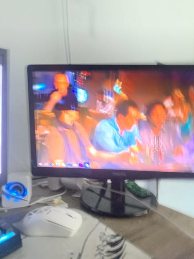 The image displays a computer monitor with a colorful, abstract image on its screen, possibly a digital artwork or a vibrant photograph. The screen shows blurred figures and shapes in shades of blue, orange, and purple, suggesting a lively and dynamic scene. To the right of the monitor, a white computer mouse is visible on a textured surface, which appears to be a desk or a tablecloth. A white speaker with a blue glowing light is positioned below the monitor, and further to the right, a dark object with a blue light is partially visible. In the background, behind the monitor and to the right, a greenish-black object with a textured surface, possibly a bag or a backpack, is seen. The overall setting appears to be an indoor space, likely a room or an office, given the presence of computer equipment. The lighting is somewhat dim, with the screen and the speaker providing some illumination. No specific time of day or weather conditions can be inferred. The image is somewhat blurry, making it difficult to discern fine details or identify specific individuals or objects with certainty. The scene does not convey any specific emotions or narrative. No visible text is present within the frame.