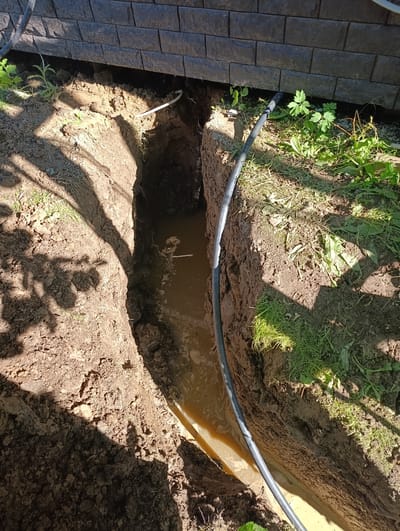 The image depicts an outdoor scene with a trench dug into the earth next to a brick wall. The trench is partially filled with murky, brown water. A black hose or pipe runs along the edge of the trench and appears to lead into or out of it. Loose soil and grass are visible on the surrounding ground. The scene appears to be located in Ryzhiki, Russia, based on the provided context. There are no people or any visible actions taking place in the image.