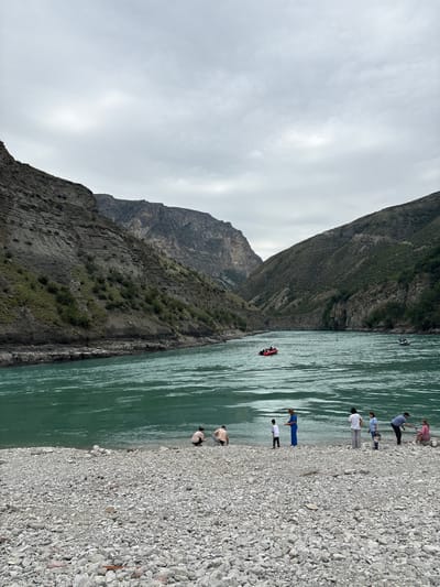 This is a wide-angle, eye-level photograph of a river valley in Chirkey, Russia, under an overcast sky. The foreground is a rocky shore composed of light-colored stones of various sizes. Several people are present on the shore. On the far left, two individuals are crouching down. Further to the right, a child stands near a woman in a blue dress. Next to them, a woman in a white top and grey pants stands with a bag, followed by a girl in a blue top and jeans. To the far right, a man is leaning towards a woman sitting on a rock.
The river, a striking turquoise color, occupies the mid-ground. It appears to be flowing, with ripples on its surface. Two boats are visible on the water: a small red raft with people in it, and a white motorboat further in the distance. The river is flanked by steep, rugged mountains on both sides. The mountainsides are covered in patches of greenery, with exposed rock faces showing distinct geological layers. The overall impression is one of a natural landscape with human activity present.