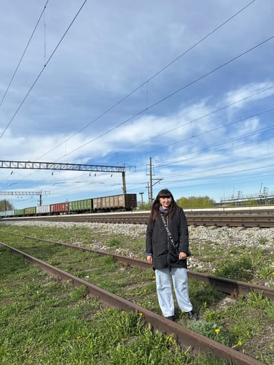 A medium shot captures a person standing on a railroad track. The individual, likely female, has long dark hair and is wearing a black jacket with repeating "GUESS" logos on the strap of a shoulder bag. Light-colored jeans and dark shoes complete the outfit.
The railroad track is overgrown with grass and weeds, interspersed with small yellow flowers. The adjacent gravel ballast supports the rails. In the background, several freight cars are visible on the track. Power lines are strung overhead, supported by metal towers. The sky is light blue with streaks of white clouds.
The image was taken outdoors on a sunny day near the city of Кечёво, Russia.