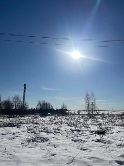 This image depicts an open, snow-covered landscape under a clear, bright blue sky in Fabriki 1 maya, Russia, on a sunny day.
The sky occupies the upper two-thirds of the frame, featuring a brilliant, undiffused sun in the upper right quadrant emitting a prominent starburst lens flare and a diagonal sunbeam. Two dark, parallel overhead power lines traverse horizontally across the upper portion of the sky, intersecting the sun's flare. A small, dark, indistinct object is visible high in the upper-left sky. A faint, wispy cloud fragment is present in the mid-left sky.
The foreground, comprising the bottom third of the image, consists of uneven, glistening snow cover with numerous sparse, dry, dark plant stalks protruding through it, casting short shadows to the left. Some areas of less dense snow reveal underlying dark ground.
In the midground, a line of leafless deciduous trees and bushes runs horizontally. To the left, a tall, slender industrial chimney or smokestack stands prominently, painted with alternating horizontal red and white stripes, its top flat. Behind the line of trees, several low-rise buildings with dark roofs are partially visible, suggesting a distant settlement or industrial area. Two slender, bare trees with light-colored bark stand slightly right of center in the midground.
The background shows a flat horizon line with a gentle rise to the right. A faint line, possibly railway tracks or distant power lines, is visible stretching horizontally across the far right background. No people or animals are present.