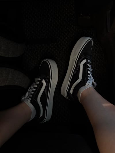 The image shows a pair of black Vans Old Skool sneakers with white laces and a white side stripe, worn by a person whose legs are visible. The shoes are on a dark, textured surface, likely a car's floor mat. The lighting is dim, suggesting an indoor or nighttime setting. The specific location in Charentsavan, Armenia, cannot be determined from the visual information provided.
