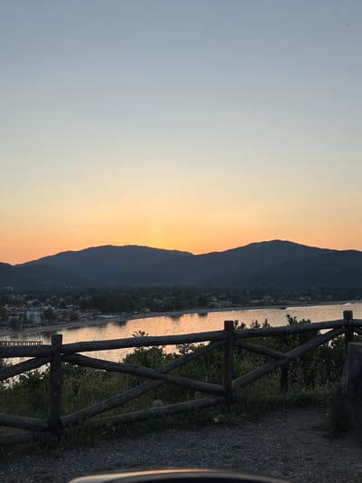 The image captures a sunset view of Olympiada, Greece. In the foreground, a rustic wooden fence runs along a path. Behind the fence, a body of water reflects the warm hues of the setting sun. Across the water, silhouetted mountains define the horizon. Nestled at the base of the mountains, a town with buildings can be distinguished. The sky transitions from a pale blue to a vibrant orange and yellow gradient near the horizon.