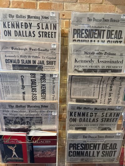 A display of historical newspapers commemorates President Kennedy's assassination. Headlines proclaim "President Dead", "Kennedy Assassinated", and "Kennedy Slain on Dallas Street". Articles detail Johnson's succession, Oswald's capture and death, and the nation's grief. Front pages from the Dallas Morning News, Fort Worth Press, New York Herald Tribune, and Pittsburgh Post-Gazette are featured. Some editions include coverage of the funeral and Kennedy's burial at Arlington Cemetery. Copies of "Legacies" books are also available.