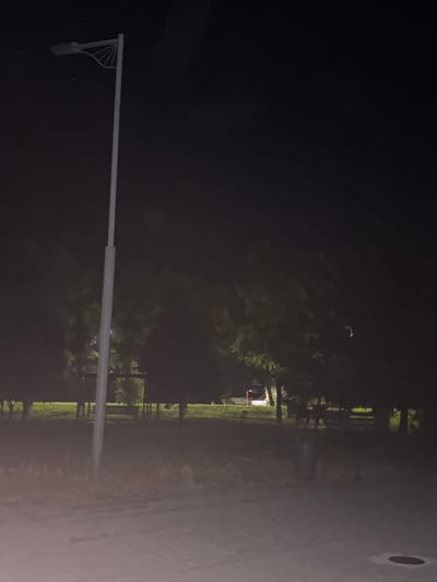 The image depicts a dimly lit scene at night, likely in a park or outdoor public space in Egorlykskaya, Russia. The primary object in the foreground is a tall, white lamppost, its arm extending horizontally across the frame. Attached to the lamppost is a light fixture, casting a faint illumination downwards. The background is dominated by dark silhouettes of trees, with hints of greenery visible. In the distance, a building with a lit window is faintly discernible, suggesting some human presence. The ground appears to be paved, possibly a walkway or road, with a faint texture visible in the lighter areas of the image. The overall atmosphere is quiet and somewhat isolated, typical of a nighttime setting in a less urbanized area. The lighting suggests it is nighttime, and the absence of harsh shadows or significant atmospheric disturbances hints at fair weather conditions. There is no visible text in the image.