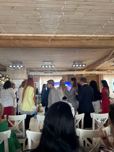 The image shows the interior of a room, likely a venue for an event, in Lilaste, Latvia. The walls are paneled with light-colored wood, adorned with strings of lights. People are seated in white chairs, facing a central area. They are dressed in a variety of formal and semi-formal attire. Some individuals appear to be looking at a screen or stage. Decorations include balloons.