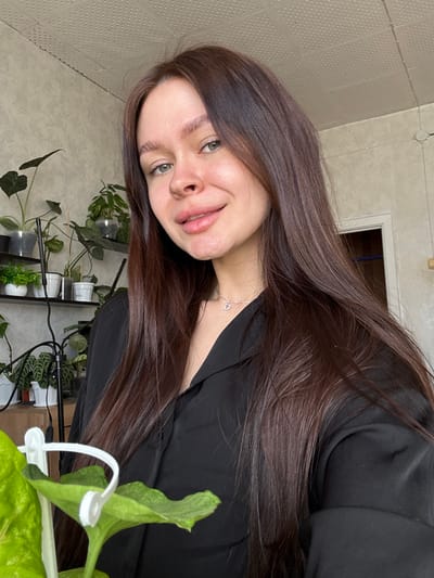 A young woman with long, dark brown hair is positioned in the foreground of the image, facing the camera at an angle. She has fair skin and her eyes are green. She is wearing a black top and a delicate silver necklace. Behind her, a collection of potted plants is visible on a shelf, suggesting an indoor environment. The background wall has a textured pattern. The presence of a plant in the immediate foreground partially obscures the view. There is nothing in the image to indicate this is from Dubrova, Russia.