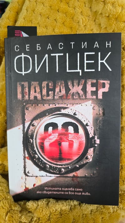 A photograph captures a paperback book resting on a textured, mustard-yellow fabric surface. The book cover is predominantly dark grey or black with white and red text and imagery. The top of the cover displays the author's name in white Cyrillic script: "СЕБАСТИАН ФИТЦЕК" (SEBASTIAN FITZEK). Below, the title "ПАСАЖЕР" (PASAZHER), meaning "PASSENGER", is rendered in distressed, textured red-brown letters.
The central artwork on the cover depicts a circular, metallic porthole-like structure, heavily distressed with visible rivets, rust, and possible blood splatters. Inside the porthole, a dark red, viscous-looking liquid or reflection partially obscures what appear to be two bold, white numeric characters, possibly "28" or "23". The lower portion of the cover features a white tagline in Cyrillic: "Истината оцелява само ако свидетелите са все още живи." (The truth survives only if the witnesses are still alive.)
A small portion of a black bookmark, featuring a white and pink stylized logo, is visible protruding from the top left edge of the book. The book is angled slightly to the right, and the fabric background exhibits a deep, irregular waffle-like texture. No identifiable persons, actions, or specific geographical landmarks are present in the image. The image does not depict the city of Wilhelmshaven, Germany, or any other specific external location; the scene is consistent with an indoor setting.