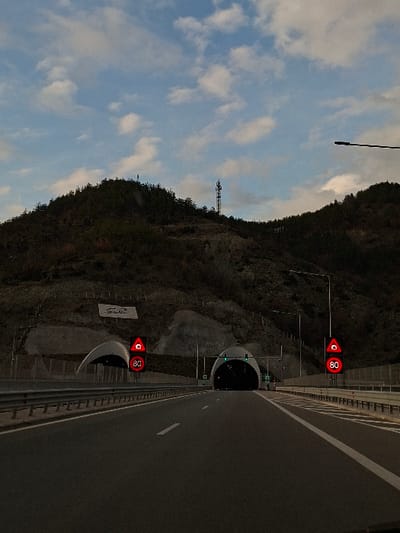 The scene depicts a multi-lane asphalt highway on the approach to the Jeleznitsa Tunnel, located within Jeleznitsa, Bulgaria. The road, featuring white lane markings and metal guardrails on both sides, leads directly into two distinct tunnel bores cut into a steep, tree-covered mountain slope.

The primary tunnel entrance is centered to the right, presenting a dark arched opening with visible green traffic lights indicating passage. To the left, a secondary, smaller arched structure or bore is visible. Positioned directly before both tunnel entrances are vertical poles supporting circular red illuminated traffic signs. Each sign displays a black warning triangle with an exclamation mark at the top, and below it, the number "80" in white against a black circle with a red border, indicating a speed limit.

The mountainous background is densely covered with trees and features exposed rock faces. On the left slope, above the smaller tunnel entrance, a large white sign with a stylized dark logo or text is affixed. Near the mountain's summit, a tall telecommunication tower with multiple antennae is visible against a partly cloudy sky. The sky displays patches of light blue with scattered white clouds. No individuals or other vehicles are visible in the immediate foreground. The overall lighting suggests either early morning or late afternoon.