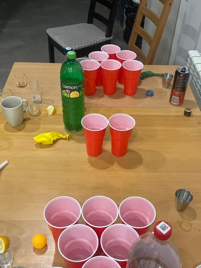 The scene is an indoor setting in Заозерное Снт, Russia, captured from an elevated perspective looking down onto a light brown wooden table. No individuals are present.
The table features various items. In the upper half, a large, partially full green plastic bottle of "Laimon fresh" beverage with a green cap stands prominently. To its left, six clear glass shot glasses/small tumblers are arranged, one containing a small amount of amber liquid and another holding a yellow lemon slice. A light grey ceramic mug is also present. Ten red plastic cups are arranged in a 4-3-2-1 triangle formation. To the right of this arrangement, a deflated green balloon, a small silver metal jigger, an empty and slightly crushed "BURN energy drink" can, two blue plastic bottle caps, and a small dark brown bottle cap are visible. A white radiator is partially seen in the upper right background.
In the lower half of the table, two standing red plastic cups are positioned. Further down, another set of ten red plastic cups is arranged in a distinct 4-3-2-1 triangle formation. An orange ping-pong ball rests near these cups. A partially visible brown glass bottle with a red cap and a white QR code sticker is at the bottom right, next to another small silver metal jigger. A yellow lemon slice is placed on a clear shot glass at the bottom left. A deflated yellow balloon lies near the table's center-left. A white cylindrical object, possibly a pen, is partially visible at the far left edge. The background reveals a dark floor, a black chair with a striped cushion, and parts of a light wooden chair.