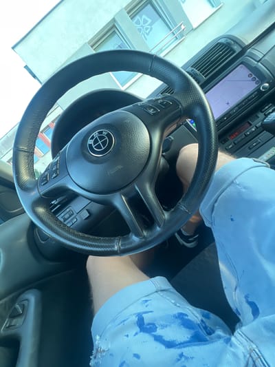 The image shows a close-up view of the interior of a car, likely a BMW, as indicated by the logo on the steering wheel. The driver's legs are visible, wearing light blue denim shorts with paint splatters. The steering wheel is black leather, and the dashboard features a navigation screen displaying a map, air vents, and climate controls. The exterior environment suggests a bright, sunny day with a building facade visible through the car windows, featuring multiple windows and light-colored cladding. The presence of the building facade and the specific architectural style point to a location in Rumia, Poland, specifically near a modern residential or commercial structure.