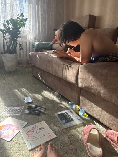 A person with wet dark hair is sitting on a beige sofa, drawing with markers on a piece of paper placed on a patterned carpet. A bottle of water, a package of markers, and slippers are nearby. A small mirror and several individual markers are also on the carpet. The person is holding a thin object, possibly a cigarette or drawing tool. A potted green plant is visible near a window covered with a sheer curtain. There is artwork present on the paper, including drawings of cats and an anime-style face. The OCR text suggests the package contains "markers" and phrases like "I want to take you home." The location is not specified.