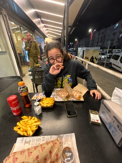 The image captures a nighttime scene at an outdoor eatery, likely in Kiryat Malakhi, Israel. The primary subject is a person with curly hair and glasses, wearing a dark long-sleeved shirt, who is seated at a black table. They are looking towards the camera, holding a single French fry, and appear to be smiling or making a playful expression.
The table is laden with food and drinks. Two items resembling shawarma or wraps are present, both wrapped in paper featuring the "MEXICANO" brand. Two portions of French fries are served in paper trays, along with a small container of dark sauce. Beverages include a red Coca-Cola can labeled "CLASSIC" and an orange-capped bottle of "fuze tea." A pack of red Marlboro cigarettes and a black smartphone are also on the table. A silver napkin dispenser bears the Hebrew text "בְּתַאֲבוֹן," meaning "bon appétit."
In the background, the scene is dark, indicating it is night, with artificial lighting from streetlights and the eatery illuminating the area. Buildings and parked cars line what appears to be a city street. Reflections on a glass surface show another person in the distance. A sign with Hebrew text and a graphic of silhouetted figures is visible further down the street. The overall ambiance is casual and suggests a late-night meal or snack enjoyed outdoors.