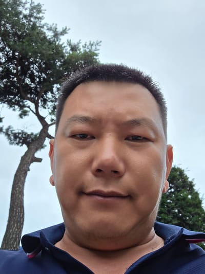 This is a vertical, close-up selfie of a person's face. The subject is a person with short, dark hair and light brown skin. They are wearing a dark blue collared shirt. The person's face takes up most of the frame, with the background consisting of a cloudy, bright sky and the branches of a large tree. The tree appears to be an evergreen with dark green foliage. The image seems to have been taken outdoors during the daytime, with the overcast sky suggesting it could be a cloudy or early morning/late afternoon. The person's expression is neutral, and the overall mood of the photo is casual. There is no visible text in the image. The location context provided is Soseok-ri, South Korea.