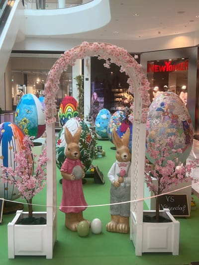The image depicts an indoor Easter-themed decorative display located within a multi-level shopping mall in Vösendorf, Austria. The central feature is a white arched trellis, adorned with artificial pink cherry blossoms, framing a pair of anthropomorphic rabbit statues. The rabbit on the left, rendered in a brownish hue, wears a pink dress and holds a grey egg. The rabbit on the right, also brownish, is dressed in a white shirt with grey suspender pants and a pink bow tie, holding a pink rose. Two smaller, solid-colored eggs (one light green, one white) rest on the green floor covering between the statues.
The display area, covered in an artificial green surface, is populated by multiple large, egg-shaped sculptures, each uniquely painted. Examples include designs featuring Winnie the Pooh and a bee, Hello Kitty, abstract rainbow patterns, various cartoon animals, and floral motifs. Two small white planter boxes, each containing an artificial cherry blossom tree, flank the base of the arch. A light-colored ribbon or string horizontally traverses the foreground of the display. A small blackboard sign, partially visible near the right planter, bears the white chalk text "VS Vösendorf".
In the background, the architecture of a modern shopping center is evident, with expansive glass storefronts. A "NEW YORKER" store sign in bright red is prominently displayed on the right. Reflections in the glass storefronts reveal further retail interiors, mannequins, and indistinct figures, including a female figure visible through a distant storefront. Overhead lighting and curved white structural elements of the mall are also visible.