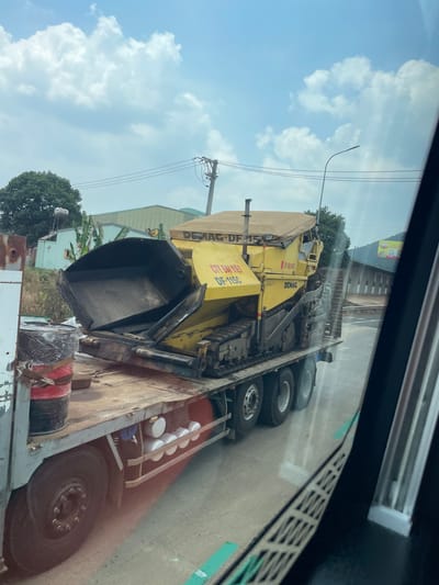 An asphalt paver, branded DEMAG, is transported on a flatbed truck in what appears to be Ia Tor, Vietnam. The truck's bed also carries several cans of paint or similar material. The sky is partly cloudy, and the view is partially obstructed by the interior of a vehicle, presumably where the photo was taken. A power pole is visible outside the vehicle, as well as buildings and some vegetation.