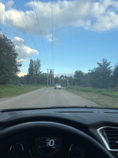 The image depicts a forward-facing view from the interior of a vehicle on a paved road in Scherwiller, France. The vehicle's dark dashboard and steering wheel occupy the lower foreground, with a digital display showing "50." The asphalt road extends into the midground, gently curving to the left. A light-colored station wagon or hatchback is visible driving away from the viewer in the middle of the road. To the right of the road, a red and white 'Give Way' (yield) sign is prominently displayed.

Overhead, multiple power lines stretch across a blue sky with scattered white cumulus clouds. These lines are supported by several utility poles, visible further down the road, constructed of concrete or metal. Extensive green foliage and trees line both sides of the road, forming a dense natural border. On the left side of the road, off the pavement in a grassy area, a dark vehicle is partially obscured. In the distant background to the right, a white recreational vehicle (camper van) is visible on what appears to be a raised embankment. No individuals are depicted.