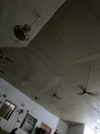 The image shows the ceiling and walls of a space, possibly a room or an outdoor area. The ceiling is paneled and has several ceiling fans and lights. The walls feature windows, a wall-mounted fan, and architectural details. The location is likely in Abeokuta, Nigeria. There are also glimpses of people near the bottom right corner of the image.
