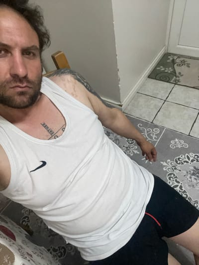 The image shows a man lying down, wearing a white tank top and black shorts. He has a tattoo on his chest and arm. The background includes a tiled floor with a rug and a white door. There is no indication of the city of Armagh, United Kingdom.