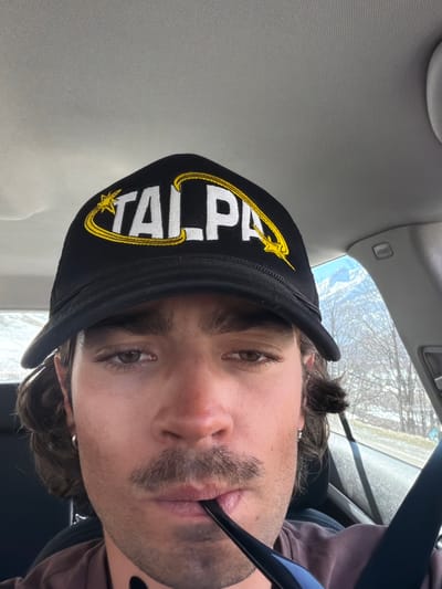 A man is wearing a black cap with white "TALPA" lettering and a yellow star/crescent design, positioned inside a car. The man is fair-skinned, with dark brown hair, light brown eyes, and a mustache/goatee. A blue object is in his mouth. The car's interior is visible, including the ceiling and airbags. Outside the window, trees and snow-covered ground suggest a winter environment. The location, Le Monêtier-les-Bains, France, is not explicitly visible in the image.