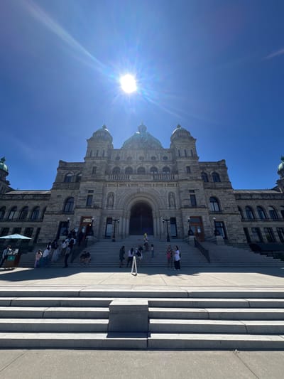 The image shows the exterior of the British Columbia Parliament Buildings, a large stone building with domes, located in Victoria, Canada. The sun is high in the sky, creating a bright glare that partially obscures the top of the image. A wide set of stone steps leads up to the main entrance of the building, where several people are seen walking and standing. Some individuals are gathered on the steps, while others are further up, approaching the entrance. The building exhibits classical architecture with arched windows and decorative stonework. To the left of the steps, there is a light blue umbrella and a cart. The foreground consists of more stone steps and a paved area. The scene is bathed in bright daylight, suggesting a clear and sunny day.