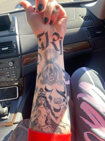 The image depicts the interior of a passenger vehicle in the city of Kruče - Kërruçi, Montenegro, under direct sunlight. The left forearm of an individual, likely a passenger, is prominently featured, held aloft with the palm facing the viewer. The forearm is extensively tattooed. On the inner wrist, black script-like characters, resembling Sanskrit or Devanagari, are visible. Above this, on the mid-forearm, is a grey stippled tattoo of an octopus. Further down the forearm, closer to the elbow, is a black outline tattoo of a multi-armed deity, possibly Kali, depicted with a crown, a third eye, exposed breasts, a protruding tongue, holding a lotus flower in one hand, and adorned with a garland of skulls. Various abstract black graphic elements surround these primary tattoos. The individual's left hand has fingernails painted with dark polish, with some minor chipping visible. The forearm is partly covered by a bright red, ribbed sleeve cuff. In the lower right of the frame, a portion of the individual's left leg is visible, clad in dark grey athletic pants or leggings featuring abstract light pink or white shapes. The vehicle's interior includes a dark dashboard with wood-grain trim, air vents, and a central console containing radio/CD player controls. Strong sunlight casts distinct shadows across the dashboard and the individual's leg.
