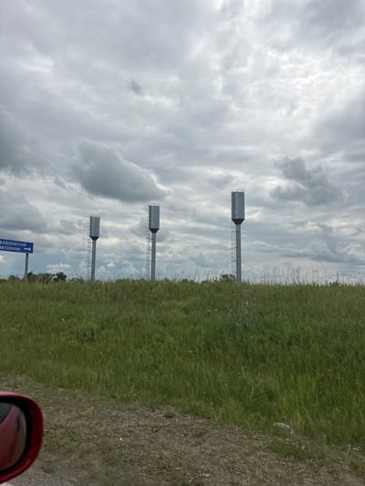 The image shows a rural landscape with three tall water towers in the background. The water towers are made of metal and have ladders running up their sides. They are positioned in a line, with the leftmost tower slightly closer to the viewer. In front of the water towers is a field of tall green grass with some sparse wildflowers. The sky is overcast with gray, heavy clouds. To the left of the water towers, a blue sign is visible with white text that appears to be in Cyrillic script, indicating directions. In the foreground, a portion of a red car's side mirror is visible on the left side of the frame, suggesting the photo was taken from a vehicle. The ground in the foreground is a dirt path. Based on the sign, the location is in the vicinity of Таштимерово, Russia.