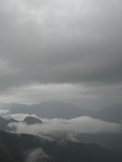 The image depicts a landscape in Rathmalkaduwa, Sri Lanka, characterized by overcast skies and shrouded mountains. Heavy cloud cover dominates the left half of the frame. On the right, a series of mountain peaks emerge through the mist. The lower slopes of these mountains are also obscured by low-lying clouds or fog. The overall scene conveys a sense of atmospheric grayness and dampness, indicative of a rainy or foggy day.