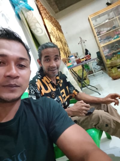 Two men are visible in the foreground. The man closest to the camera is wearing a dark green t-shirt. The other man is wearing a patterned shirt with black and yellow designs and khaki pants. He is seated on a green plastic chair. The location appears to be indoors, possibly a residential setting in Dayahlhoksukon Barat, Indonesia. A glass cabinet is in the background, and a small folding table with various items is placed next to a wall.