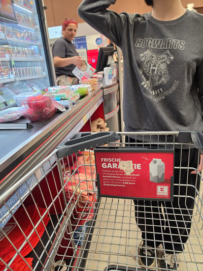 The image captures a close-up view of a shopping cart in a supermarket, with a person's torso and arm visible in the upper left, wearing a gray sweatshirt featuring the Hogwarts crest and "HOGWARTS" text. The cart itself is the primary focus, filled with various items. A prominent red sign is attached to the side of the cart, displaying German text. The sign's text, "FRISCHE GARANTIE" (Freshness Guarantee), explains that if a product is found past its best-before date, the store will provide a 2.50 Euro voucher. The Kaufland logo is also visible on this sign.

In the background, another person with red hair is seen in the reflection of a refrigerated display case, holding a product and looking towards the cart. The display cases appear to be stocked with food items. The overall scene suggests a shopping trip in a German supermarket, likely during daytime hours given the lighting. The atmosphere seems casual, as one might expect during a routine shopping excursion. Red reusable shopping bags are visible at the bottom of the cart, and some produce, like red apples in a net bag, are also present in the cart and the display. The text "TSCHÜSS EIN WEG" (Goodbye One Way) is partially visible on a blue label near the bottom of the cart, possibly related to recycling or disposal.