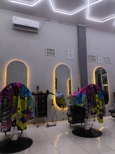 The image shows the interior of a barber shop in Miruektaman, Indonesia. The shop is brightly lit with a modern, geometric ceiling light installation. The walls are a light gray, and there are several framed certificates displayed on the wall. Two barber chairs, draped with colorful capes featuring a graffiti-like design, are visible in the foreground. Behind them, a silver trolley cart with black drawers holds barbering supplies, including a hairdryer and spray bottles. Two large, arched mirrors with yellow backlighting are positioned on the wall, one of which has a clock decal. The floor is tiled with white squares that reflect the ambient light. An air conditioning unit is mounted on the wall. The scene is static and depicts an unoccupied barber shop, suggesting it is either closed or the staff and clients are out of frame.