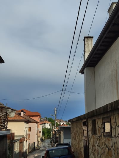 The image depicts a street scene in Pletena, Bulgaria, under a dramatic sky with dark clouds and hints of blue. The perspective is looking uphill on a narrow street lined with residential buildings. In the foreground, to the right, is a low stone-faced building with two windows, one reflecting light. To the left, a partially constructed wooden structure is visible.

The street itself appears wet, suggesting recent rain. Cars are parked along the side of the street, and a blue car is partially visible in the bottom right corner. A utility pole with multiple wires stretches across the scene. Further up the street, other houses with red-tiled roofs are visible, nestled amongst green trees. The architecture suggests a Balkan village or town. Smoke is faintly visible emanating from a chimney on one of the houses, indicating activity within the dwellings. The overall atmosphere is slightly overcast and moody, with the interplay of light and shadow from the dramatic sky. There are no visible people in the frame.
