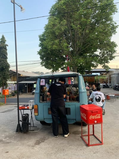The image depicts an outdoor mobile beverage stand operating in Landungsari, Indonesia. In the foreground, a person in a black t-shirt and dark pants stands with their back to the viewer, actively preparing drinks within the open rear of a light blue van, which is configured as a service counter with bottles and various containers. To the immediate right of the van, a red Coca-Cola branded cooler is elevated on a matching red metal stand. Further right, two individuals are seated, one wearing a white t-shirt with an 8-ball logo, looking towards the vendor. To the left of the van, a small dark table supports a portable gas burner, with a black trash bag positioned beneath it. A tall, black metal street light pole extends vertically on the far left. The background features a large, leafy green tree, from which a red and white flag is hanging. Beyond the tree, various buildings and structures, along with several parked motorcycles, are visible. The ground is a light-colored paved surface, and overhead utility wires crisscross the sky. The scene is illuminated by natural daylight.