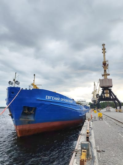 A blue cargo ship named "EUGENII ORLOVSKII" is docked at a quay in Shal'skii, Russia, under a cloudy sky. The ship's hull is painted blue with a red waterline. The quay is made of gravel and is lined with industrial equipment, including large yellow cranes. There are no people visible in the image. The water is dark and choppy. The scene suggests a working port environment, possibly for cargo loading or unloading.