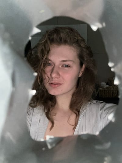 An individual, identified as a female, is depicted looking directly into the camera through an irregular, light-colored foreground aperture. Her face is centered, featuring fair skin, light-colored eyes, and shoulder-length wavy brown hair. She is wearing a partially unbuttoned, light-striped collared shirt. The background is dimly lit and out of focus, revealing a dark wall and a woven container holding light-colored objects to the right. The scene is illuminated by an unspecified internal light source. The location is Tashevka, Russia.
